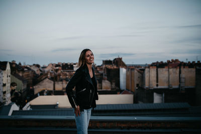 Smiling young woman standing against cityscape during sunset