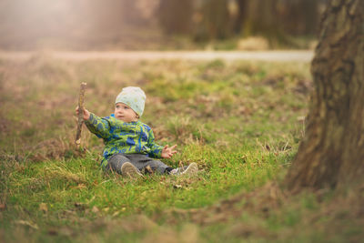 Full length of cute baby boy holding stick while sitting on grass at park