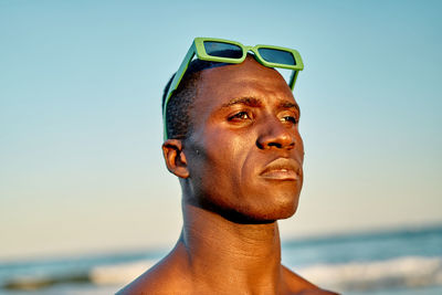 Serious african american male with green sunglasses on head looking away against sea and cloudless sundown sky