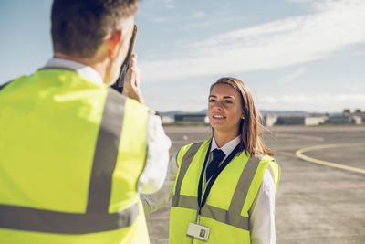 Rear view of male engineer showing airplane parts to female trainee while standing against blue sky on airport runway