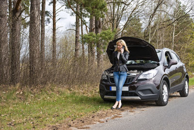 Full length of fashionable young woman standing by breakdown car on roadside