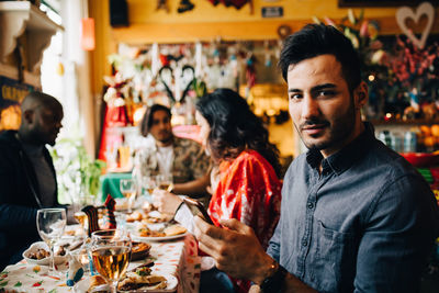 Portrait of confident young man holding smart phone while sitting with friends at table in restaurant during dinner part