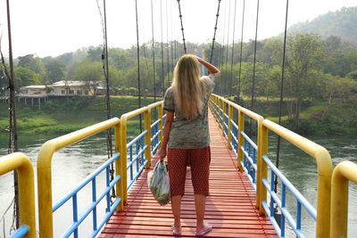 Rear view of woman standing on footbridge over lake against trees