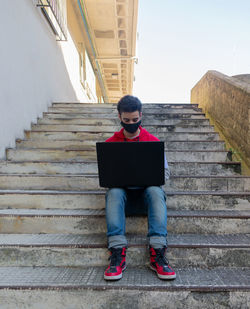 Young caucasian man dressing casual working on his laptop while sitting on some stairs with a mask