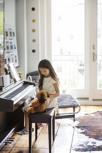 Girl sitting with teddy bear by piano at home