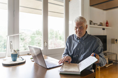 Senior man sitting at home in front of laptop turning pages in notebook