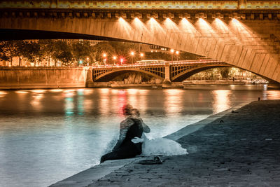 Digital composite image of bride and groom siting by illuminated bridge over river in city at night