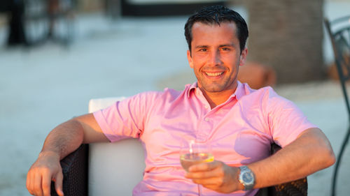 Portrait of man holding wineglass while sitting on chair at dusk