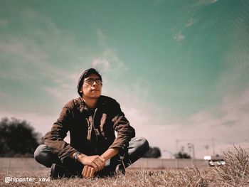 Portrait of young man sitting on field against sky