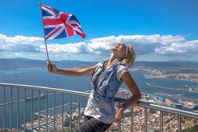 Happy woman holding british flag while leaning on railing against cityscape