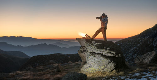 Photographer photographing by bonfire on rock against clear sky during sunset