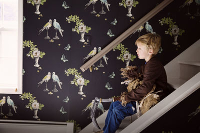 Side view of boy holding costume while sitting on staircase at home