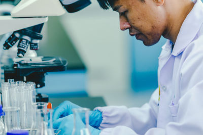 Asian man lab technician in protective glasses and gloves sits next to a microscope in laboratory.