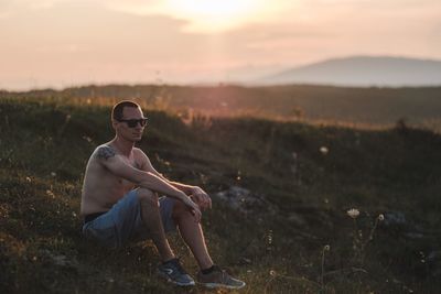 Shirtless young man sitting on field against sky during sunset