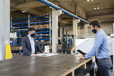 Businessman wearing face mask using digital tablet while standing at social distance with colleague in factory