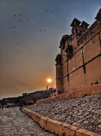 Birds flying over buildings against sky during sunset