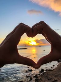 Cropped hands of person making heart shape at beach during sunset