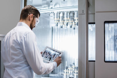 Man wearing lab coat and safety goggles at machine in factory looking at tablet