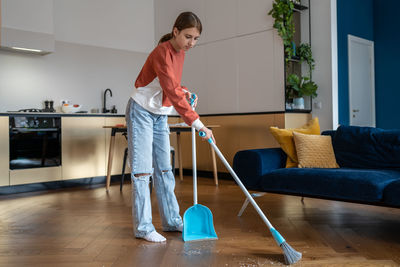 Teen girl doing chores cleaning floor in kitchen sweeping trash with broom to scoop.