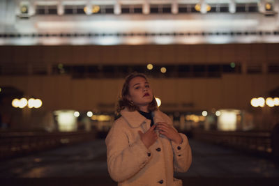 Young woman looking away standing against illuminated city at night