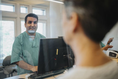 Male receptionist talking with patient through glass screen at reception desk in hospital