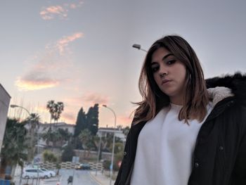 Low angle portrait of young woman standing against sky during sunset