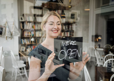 Smiling woman in a cafe attaching open sign to glass pane