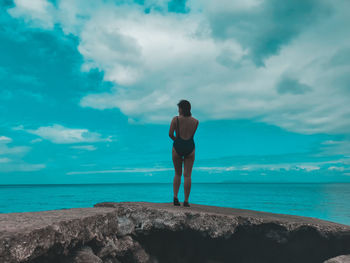 Rear view of woman in swimwear standing on rock by sea against sky