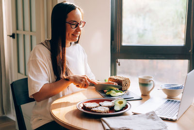 Young modern woman making healthy sandwich and video chatting on laptop at the same time.