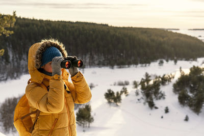 Woman in yellow looking through binoculars at birds on snow river winter forest birdwatching ecology