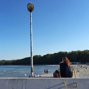 Rear view of woman sitting on retaining wall at beach against clear sky