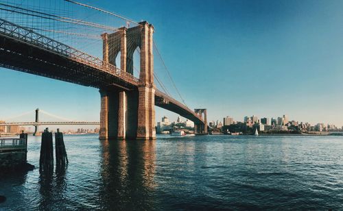 Low angle view of brooklyn bridge over east river by city against blue sky