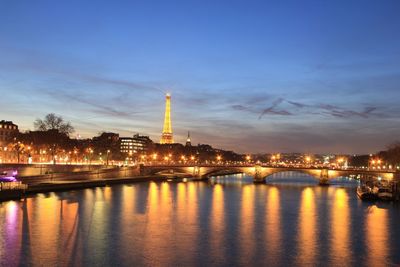 Illuminated buildings by river against sky at sunset