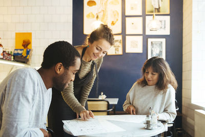 Smiling young waitress assisting customers choosing from menu at table in cafe