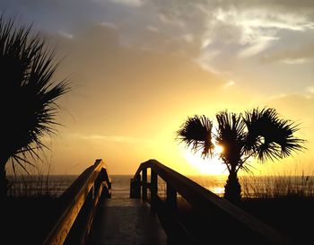 Footbridge leading towards beach against sky during sunset