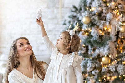 Cute girl toddler and her mom decorate the christmas tree in the house. happy family