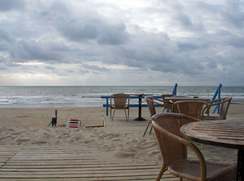 Empty chairs and table at beach against cloudy sky