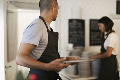 Man holding serving tray looking at female colleague standing by stack of plates in cafe