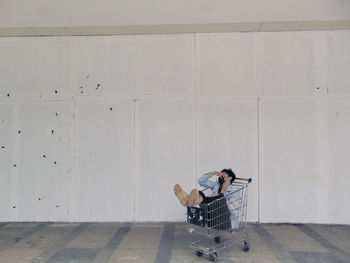 Man sitting in shopping cart against wall