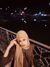 Portrait of young woman standing against railing at night