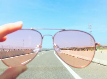 Cropped hand holding sunglasses over road against clear sky