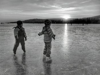 Siblings ice-skating on rink against sky during sunset