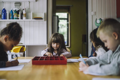 Multiracial male and female students writing on paper at table in kindergarten