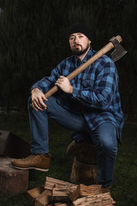 Full length portrait of lumberjack holding axe while sitting on log at forest