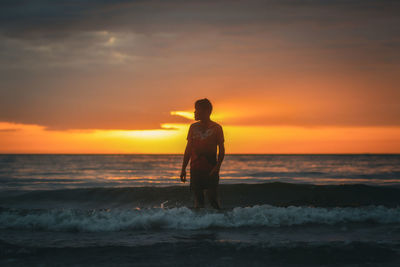 Rear view of man standing at beach against sky during sunset