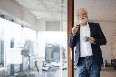 Senior businessman holding cigarette with coffee cup while leaning on glass wall at office