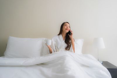 Young woman sitting on bed against wall at home