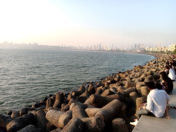 Rear view of man sitting on rock by sea against sky