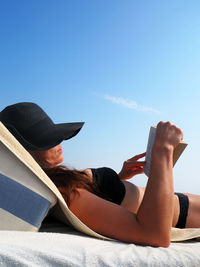 Young woman reading book while relaxing against sky
