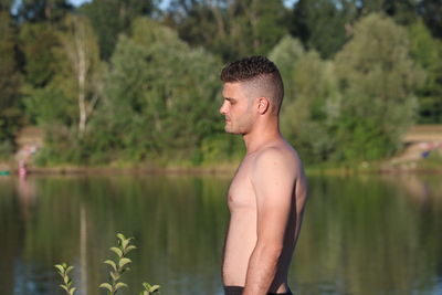 Young man looking away while standing by lake against trees